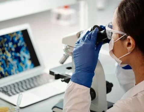 A scientist wearing a lab coat, gloves, safety goggles, and a mask is looking into a microscope. A laptop displaying a colorful image is open on the desk beside them. Laboratory equipment is visible in the background.