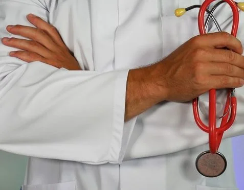 A person in a white lab coat stands with arms crossed, holding a red stethoscope.