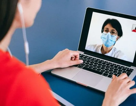 A person in a red shirt is having a video call on a laptop with another person who is wearing glasses and a face mask. Earbuds are in use, and the setting appears to be a casual environment.