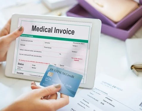 A person holding a tablet displaying a medical invoice alongside a credit card. The invoice includes personal details, status, and income fields. A purple pen is nearby, and a clipboard with documents is partially visible in the background.