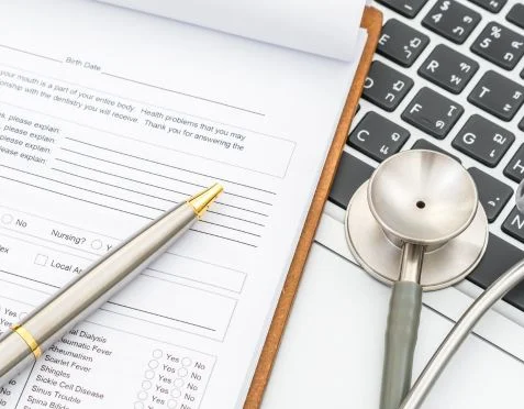 A close-up of a medical form on a clipboard with a pen resting on it. A stethoscope lies nearby on a laptop keyboard. The image conveys a healthcare or medical theme.