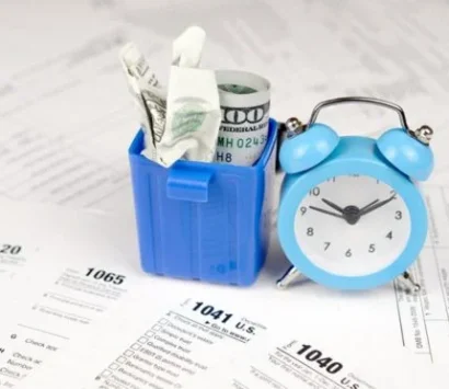 A small blue trash bin filled with crumpled dollar bills is next to a blue alarm clock. Both items are on top of various tax forms spread across the surface.