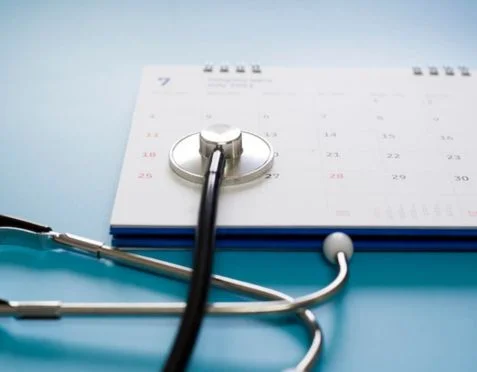 A stethoscope rests on an open calendar displaying a months grid on a light blue surface. The image suggests scheduling or planning related to medical appointments or healthcare.