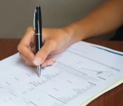 A person holding a black pen, poised to write on a document placed on a wooden surface. The document includes printed text and a visible signature line, suggesting its an official paper or form.