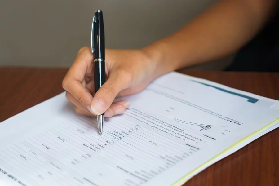 A person holding a black pen, poised to write on a document placed on a wooden surface. The document includes printed text and a visible signature line, suggesting its an official paper or form.