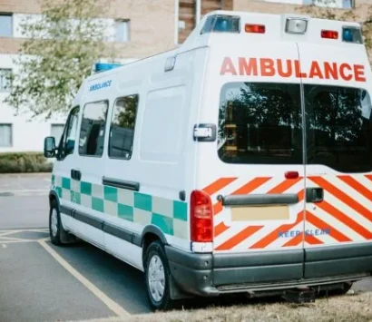 A white ambulance with red and green markings is parked in a designated area on a sunny day. The rear of the vehicle is visible, showing the word AMBULANCE in red letters above the back windows. Trees and a brick building are in the background.