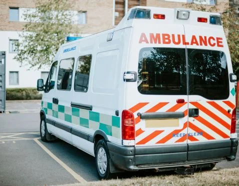 A white ambulance with red and green markings is parked in a designated area on a sunny day. The rear of the vehicle is visible, showing the word AMBULANCE in red letters above the back windows. Trees and a brick building are in the background.