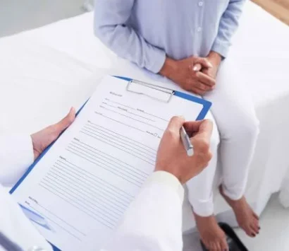 A doctor is holding a clipboard with a form, preparing to write. A patient wearing a light blue shirt and white pants sits on an examination table with hands clasped, facing the doctor. The room has a bright, clinical setting.