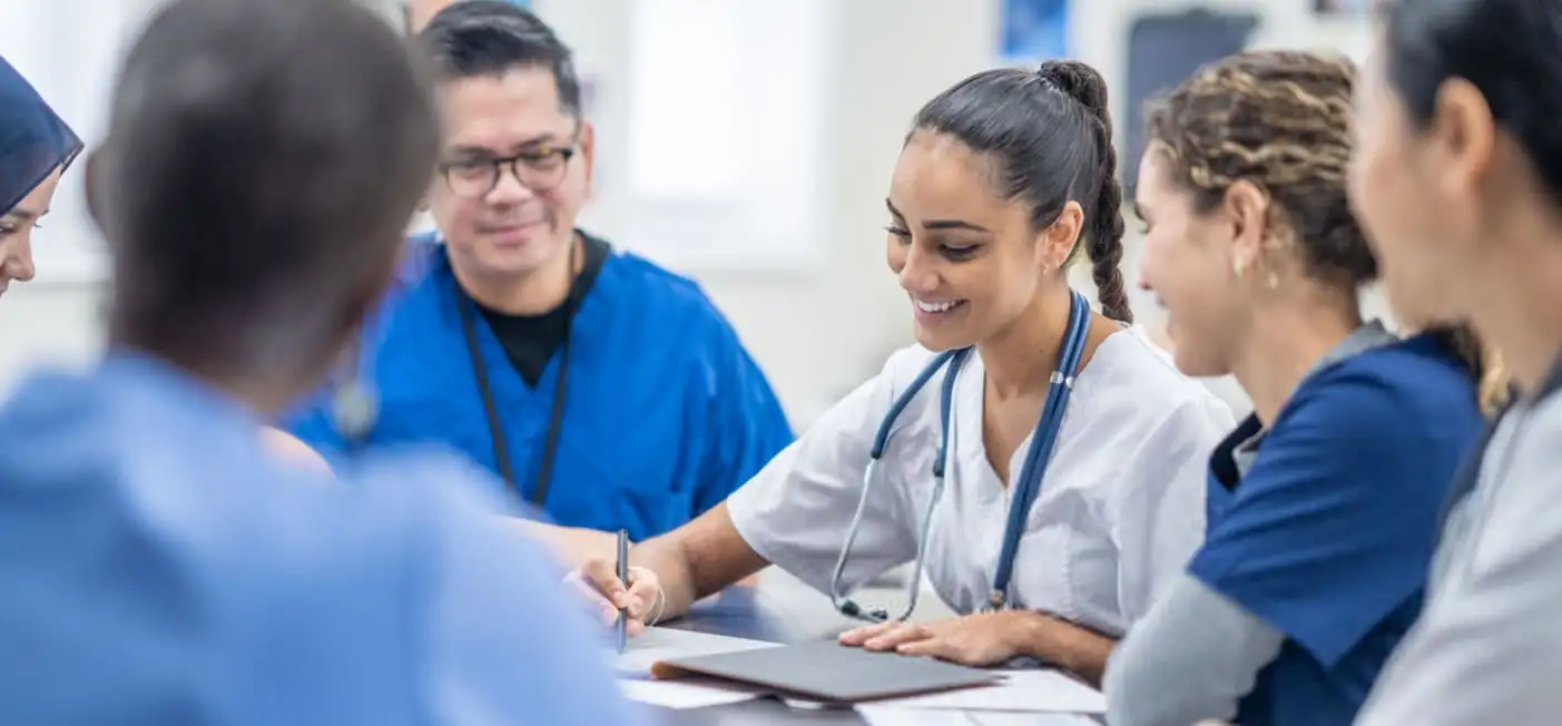 A group of healthcare professionals in scrubs sit around a table having a discussion. One person is writing on a notepad, and everyone is smiling and engaged. The setting appears to be a bright, modern medical facility.