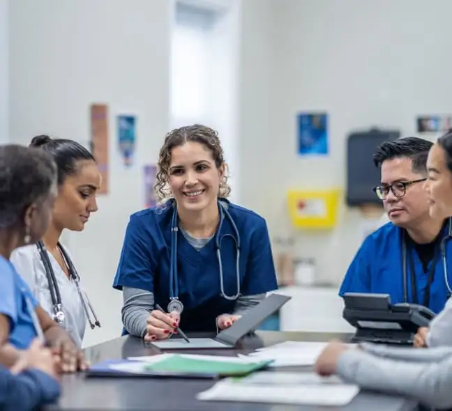 A diverse group of healthcare professionals sits around a table in a meeting room. They are wearing scrubs and have stethoscopes. One person is smiling and holding a tablet. Papers and a phone are on the table.