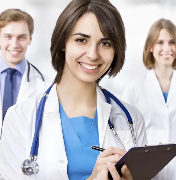 A smiling doctor with short brown hair holds a clipboard in the foreground, wearing a white coat and stethoscope. Two other doctors, a male and female, stand behind her, also dressed in medical attire, all looking towards the camera.