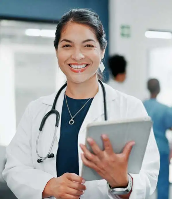 A smiling doctor wearing a white coat and stethoscope holds a tablet in a medical setting. She stands against a blurred hospital background, exuding a professional and approachable demeanor.