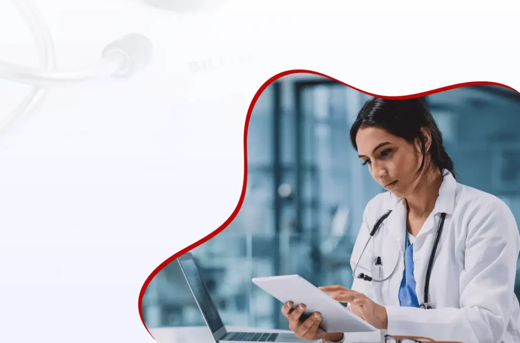 A doctor wearing a white coat and stethoscope sits at a desk, using a tablet and a laptop, focused on her work in a clinical or hospital setting.