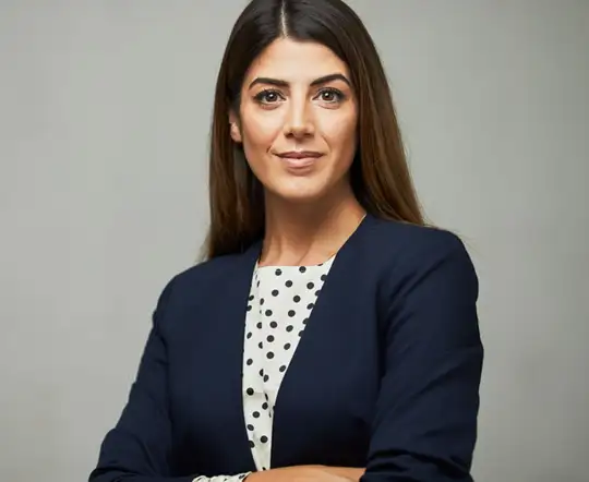 A confident woman with long brown hair wearing a navy blazer and a white blouse with black polka dots. She has a subtle smile and stands with arms crossed against a plain gray background.