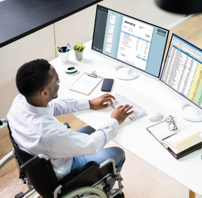 A person sitting in a wheelchair works at a desk with a computer setup. They are typing on a keyboard, with two monitors displaying an invoice and a spreadsheet. A phone, notepad, pen, and mouse are on the desk. The environment is bright and organized.