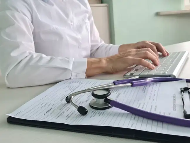 A person in a white shirt types on a keyboard. A clipboard with papers and a stethoscope rests on the desk.