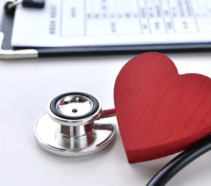 A stethoscope is placed next to a red wooden heart on a white surface. Behind them, a clipboard with medical paperwork is partially visible. The scene conveys themes of healthcare and cardiology.