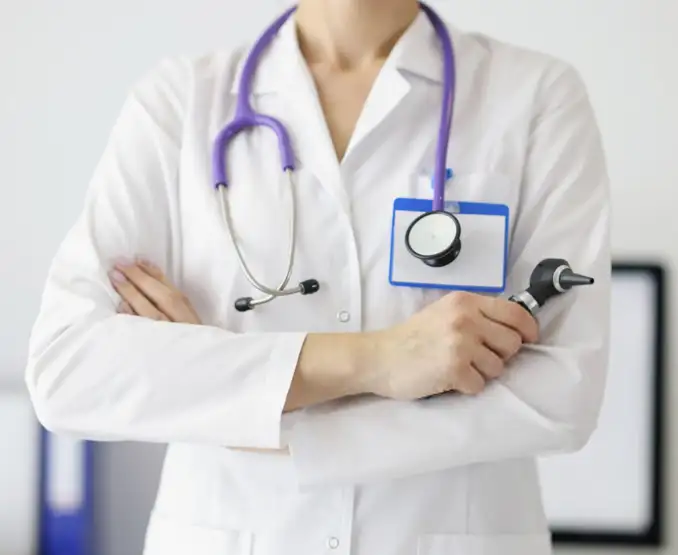 A medical professional in a white coat is shown with arms crossed. They wear a stethoscope around the neck and have a name badge on the chest. An otoscope is held in one hand, indicating a healthcare setting.