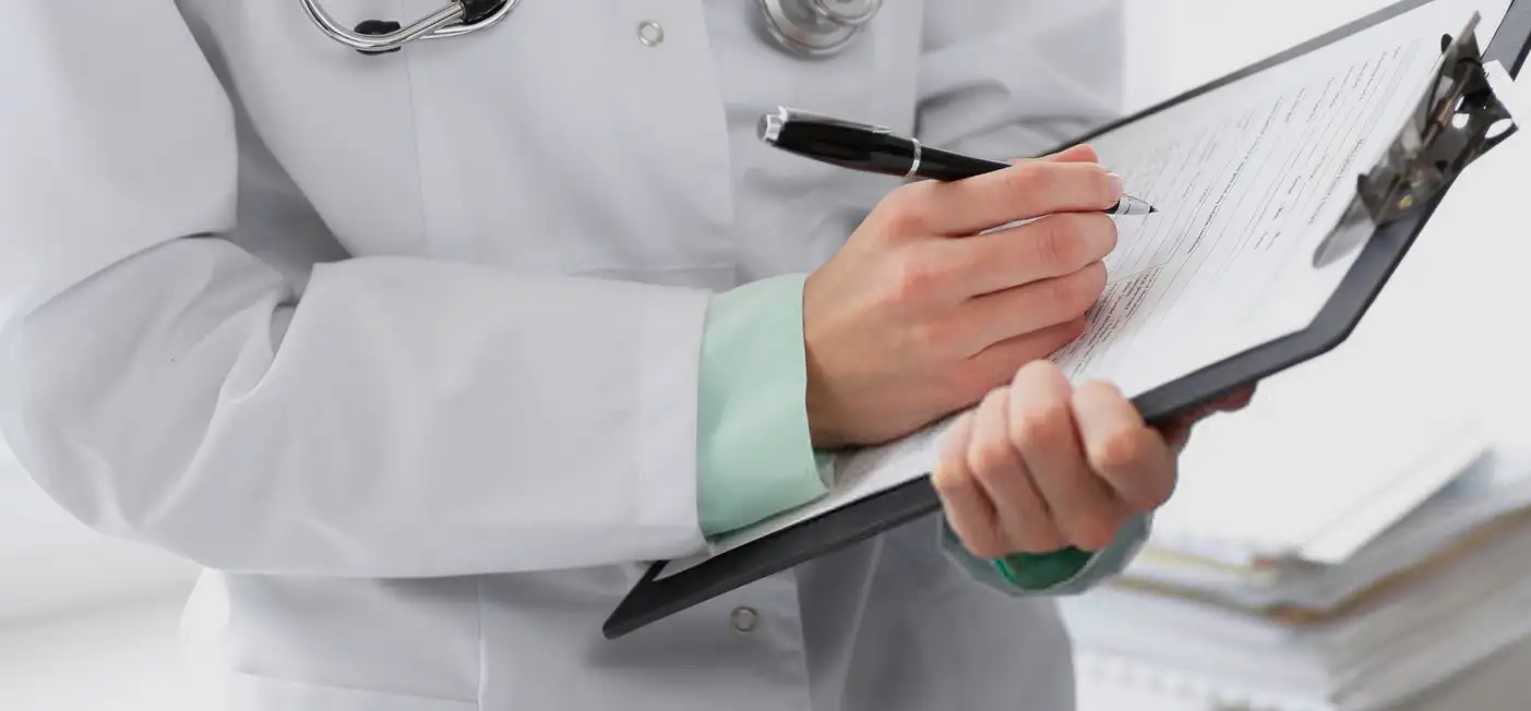 A doctor in a white coat is holding a clipboard and writing on a form with a pen. The image focuses on the doctors hands and the clipboard, with a stethoscope visible around the neck.