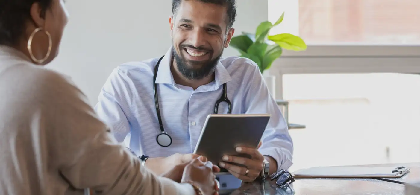 A doctor with a stethoscope around his neck is smiling and holding a tablet while speaking with a patient across the table. There is a clipboard on the table and a plant in the background.